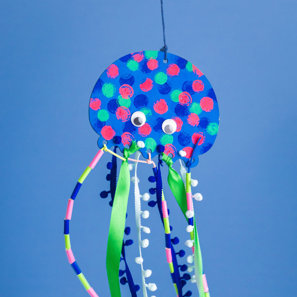 The finished Jelly! the Jellyfish project: a colorfully painted wooden jellyfish with flowing tentacles made of silk ribbon, pompom trim, twine, and straws.