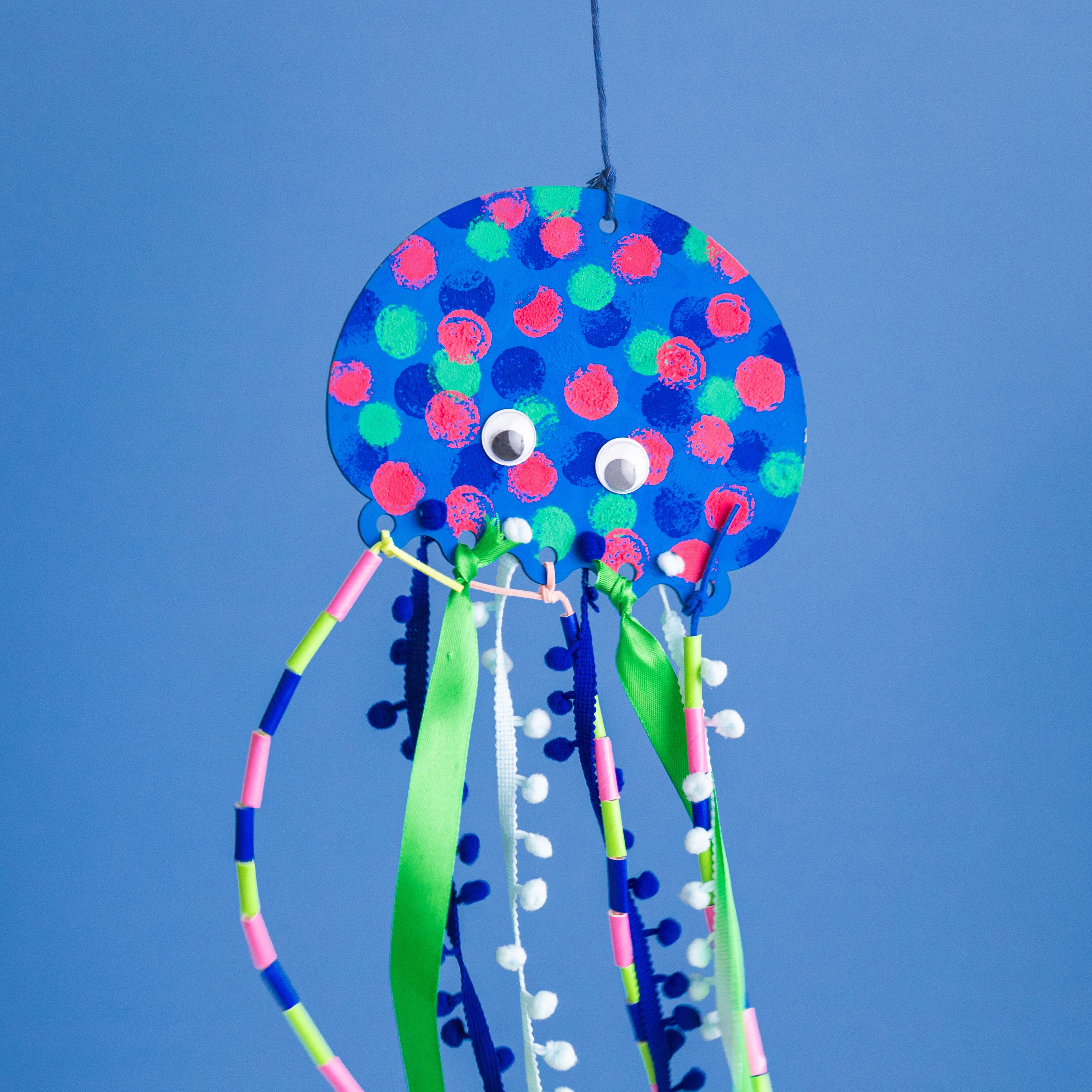 The finished Jelly! the Jellyfish project: a colorfully painted wooden jellyfish with flowing tentacles made of silk ribbon, pompom trim, twine, and straws.