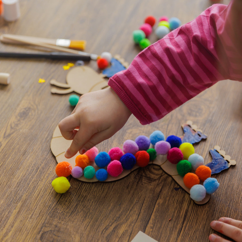 Detail of a hand using glue to attach soft, colored pompoms to the back of the wooden caterpillar.